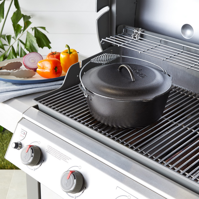 Black cast iron Dutch oven with spiral bail handle and fitted lid resting on a grill burner, surrounded by fresh vegetables, ideal for Kitchen & Food, Cookware, Cast Iron, USA Made, Best Sellers, Promo Eligible collections.