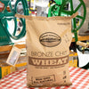 Large kraft paper bag of hard red wheat berries labeled non-GMO and all natural, placed on a checkered tablecloth with vintage grain mills in the background, ideal for Kitchen & Food and Bread Making Supplies collections.