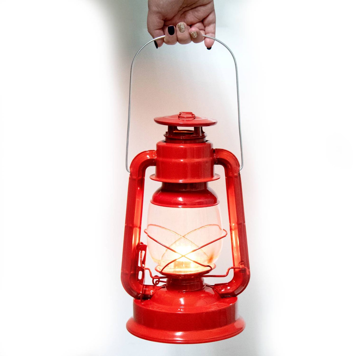 Bright red metal lantern with glass globe and lit flame, held by a hand, designed for outdoor use in farm, garden, camping, emergency, and pest control settings.
