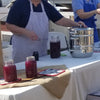 Person in striped apron operating a stainless steel steam juicer with glass lid on a white table, surrounded by jars of dark red juice, highlighting kitchen and food preparation in a casual setting.