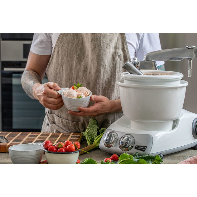 Person wearing a beige apron holding a bowl of freshly made ice cream next to a white ice cream mixer attachment on a countertop with strawberries and leafy greens, suitable for Kitchen & Food, Kitchen Gadgets, and Specialty Equipment collections.