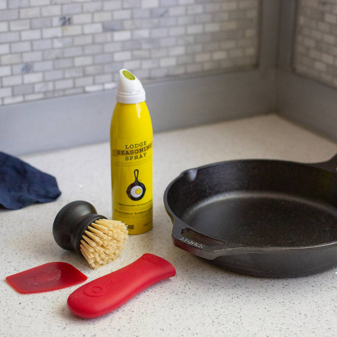 Cast iron cookware care kit featuring a yellow seasoning spray, nylon scrub brush with black handle, red silicone handle holder, and red pan scraper on a kitchen counter, ideal for Home & Decor, Kitchen & Food, Cast Iron, and Cleaning Utensils collections.