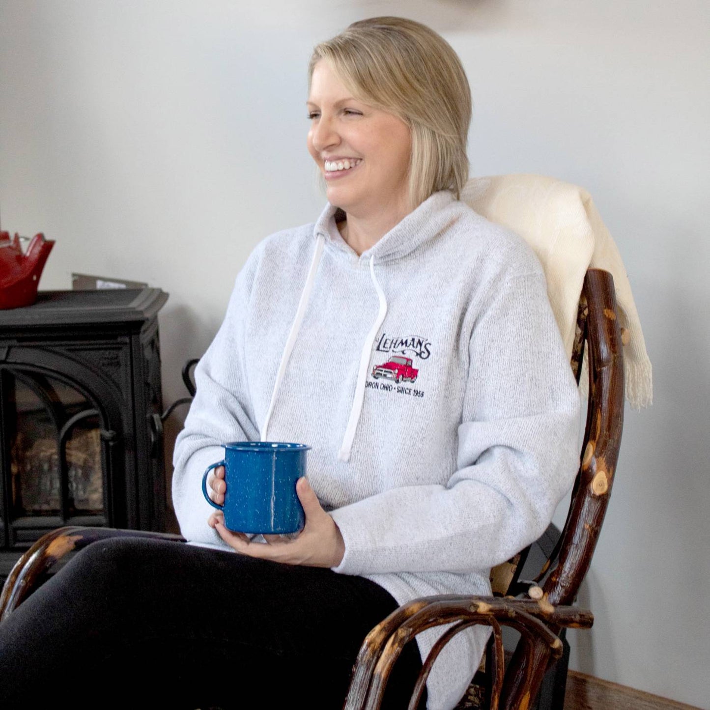 Smiling woman wearing a handcrafted light gray tunnel neck sweatshirt embroidered with a classic red truck and Lehman’s logo, sitting in a wooden chair holding a blue mug, featured in Health & Wellness, Clothing and Accessories, Best Sellers, and Promo Eligible collections.