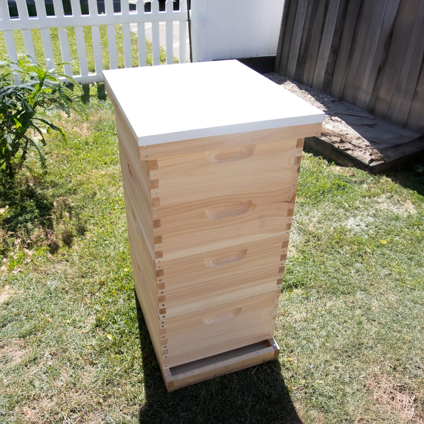 Handcrafted wooden beehive with a smooth natural finish topped by a white telescoping metal cover, set outdoors on grass near a wooden fence, Amish made, USA made, ideal for farm and garden use.