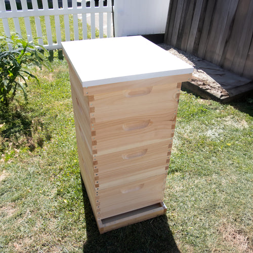 Handcrafted wooden beehive with a smooth natural finish topped by a white telescoping metal cover, set outdoors on grass near a wooden fence, Amish made, USA made, ideal for farm and garden use.