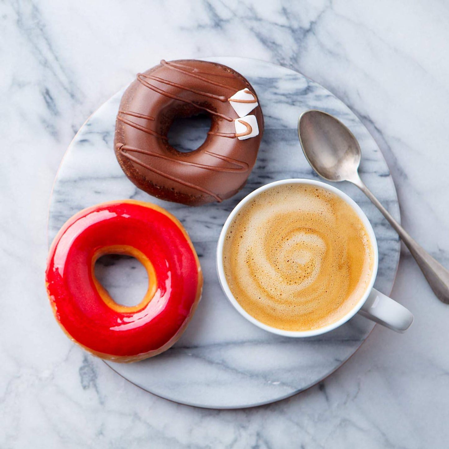 Round polished marble tray with smooth surface, holding a cup of frothy coffee, a silver spoon, and two colorful glazed donuts, ideal for stylish kitchen and dining settings.