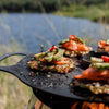 Close-up of a rustic griddle plate on an outdoor stove cooking topped potato pancakes with sliced avocado, tomato, and red chili, perfect for camping, grilling, and farm tool collections.