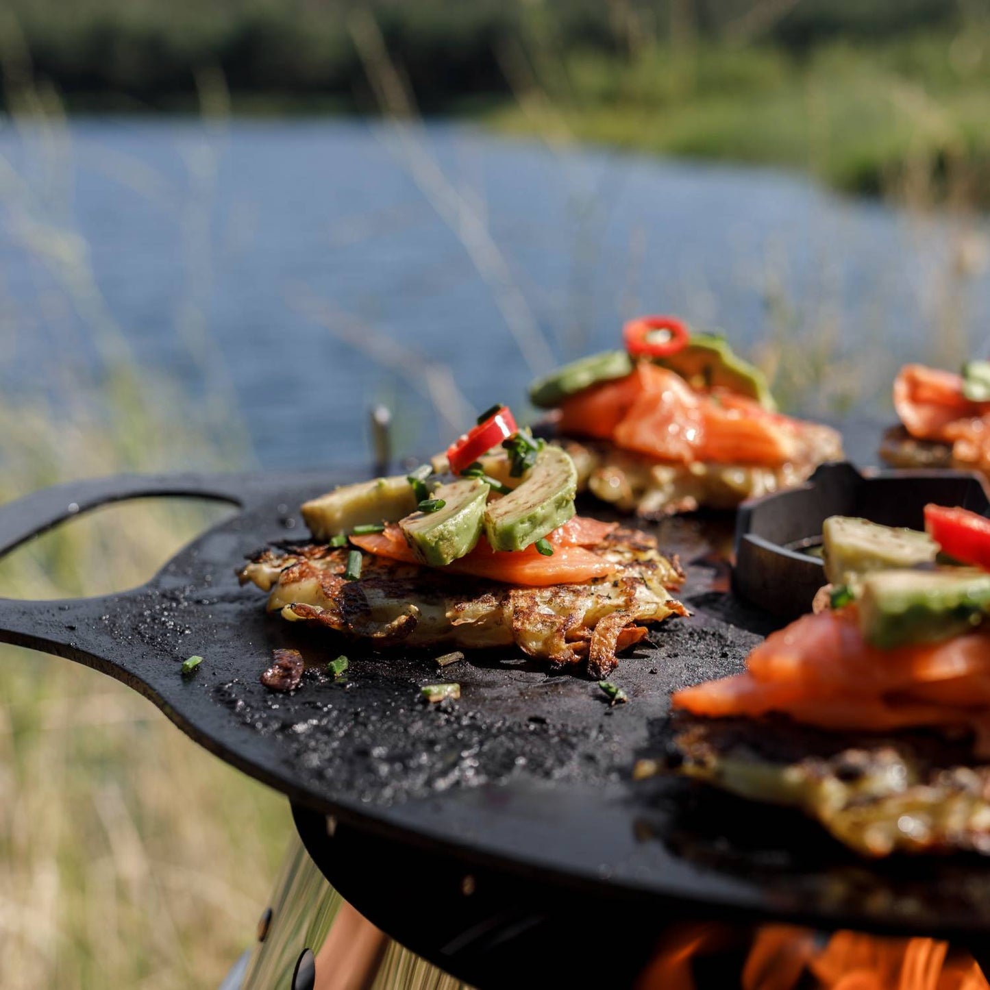 Close-up of a rustic griddle plate on an outdoor stove cooking topped potato pancakes with sliced avocado, tomato, and red chili, perfect for camping, grilling, and farm tool collections.