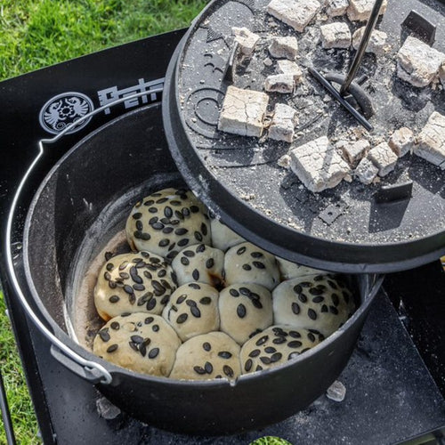 Handcrafted cast iron Dutch oven with flat base baking golden rolls topped with seeds, outdoor cooking setting featuring charcoal briquettes on embossed lid, Kitchen & Food, Cookware, Cast Iron, Best Sellers, Promo Eligible collections.