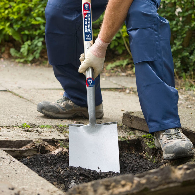 Person wearing work gloves and boots using a durable treaded digging spade to break through soil on a paved garden path, ideal for Farm, Garden & Tool, Digging and Cultivating, Lawncare, Best Sellers, and Promo Eligible collections.