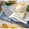 Stainless steel serrated pie server with a smooth handle resting on a striped cloth beside a lemon cream pie slice garnished with whipped cream and lemon zest, ideal for Kitchen & Food, Storage and Serving, USA Made, Best Sellers, and Promo Eligible collections.