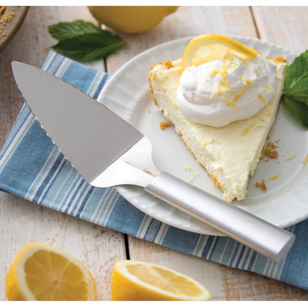 Stainless steel serrated pie server with a smooth handle resting on a striped cloth beside a lemon cream pie slice garnished with whipped cream and lemon zest, ideal for Kitchen & Food, Storage and Serving, USA Made, Best Sellers, and Promo Eligible collections.