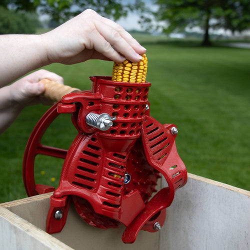 Handcrafted cast iron corn sheller in bright red with wooden handle being operated outdoors to quickly strip kernels, part of Farm, Garden & Tool and Farm Tools collections.