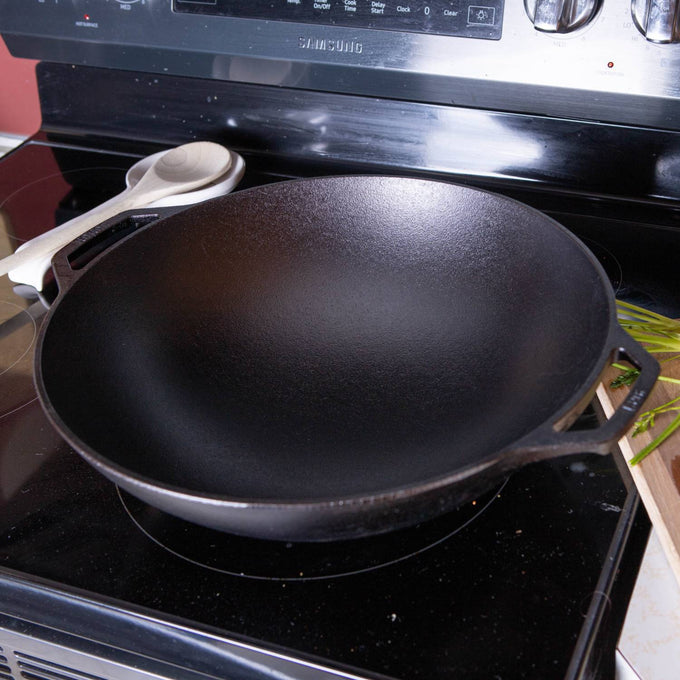 Black cast iron wok with wide, deep bowl-shaped base and dual handles, positioned on a stovetop, highlighting durable construction and ideal heat retention for Kitchen & Food, Cookware, Cast Iron, USA Made, and Best Sellers collections.
