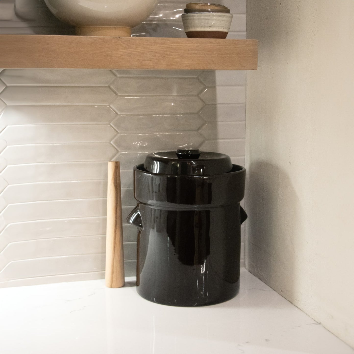 Glossy black stoneware fermenting crock with wooden stomper, set on a white countertop under a wooden shelf, handcrafted and ideal for Kitchen & Food, Food Preservation, and Fermenting collections.