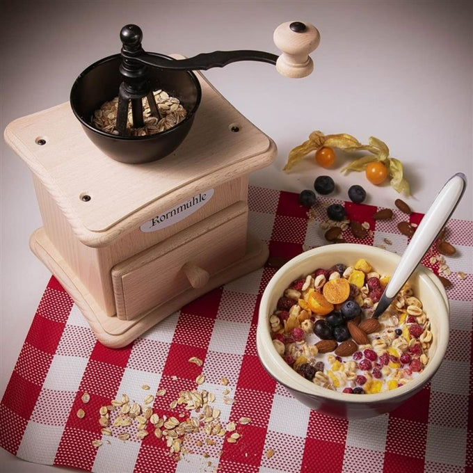 Light wooden hand-crank grain mill with a black grinding bowl filled with oats, placed next to a bowl of mixed cereal with milk and fresh berries on a red and white checkered cloth, ideal for Kitchen & Food and Bread Making Supplies collections.