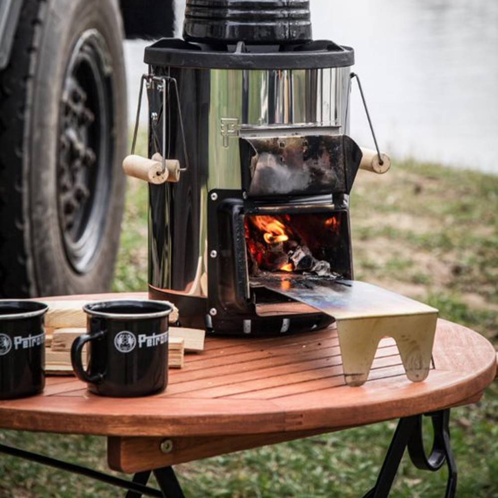 Compact portable wood stove with a polished steel body and wooden handles, shown burning firewood on a wooden outdoor table near a vehicle, ideal for Camping, Farm, Garden & Tool, and Best Sellers collections.