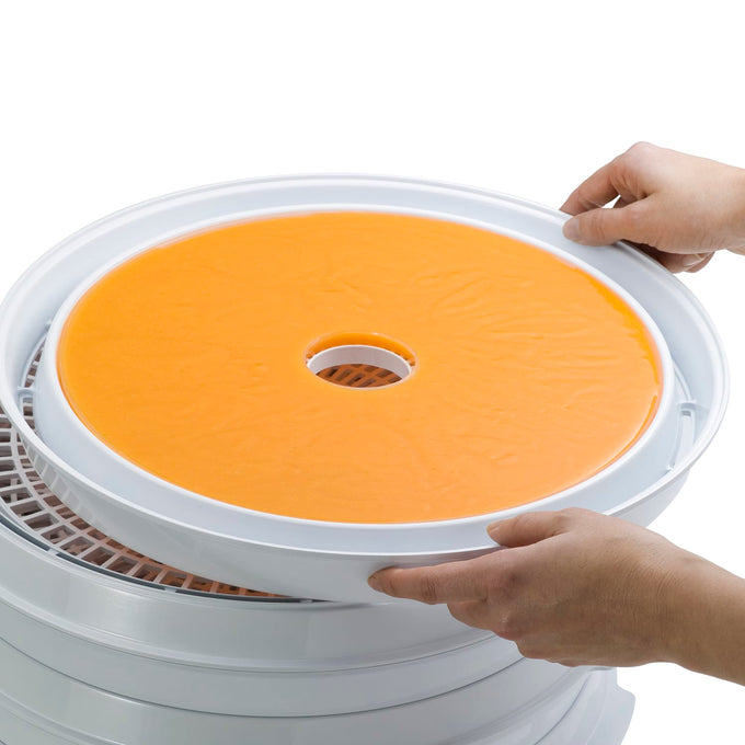 Hands placing an orange fruit roll sheet on a white dehydrator tray, shown as part of kitchen and food preservation equipment for drying and making homemade snacks.