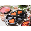 Cast iron heart-shaped mini cake pan with four cavities, featuring three baked red cakes, set on a white tabletop with pink plates, glasses, and brass candle holders, part of Kitchen & Food and Cookware collections.