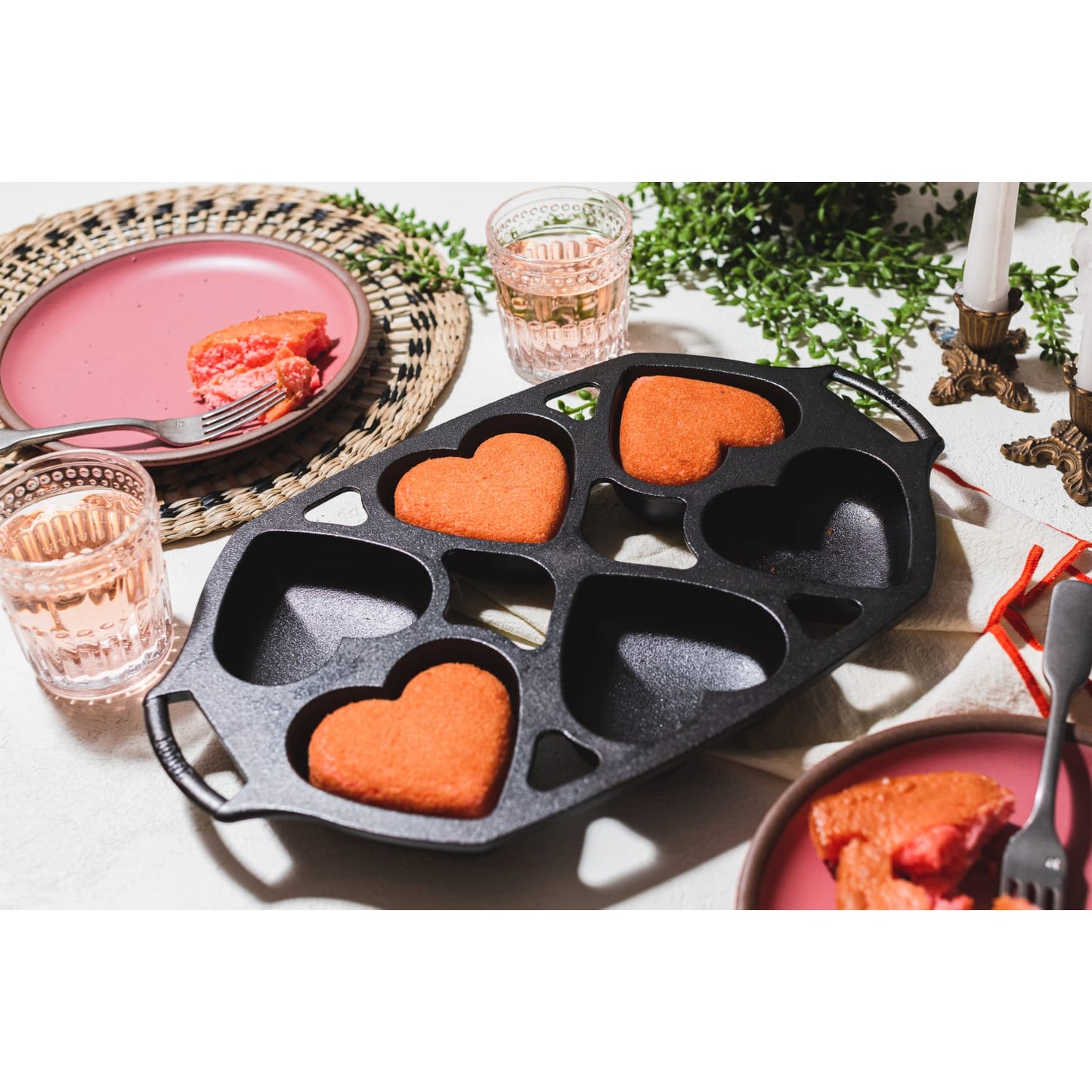 Cast iron heart-shaped mini cake pan with four cavities, featuring three baked red cakes, set on a white tabletop with pink plates, glasses, and brass candle holders, part of Kitchen & Food and Cookware collections.