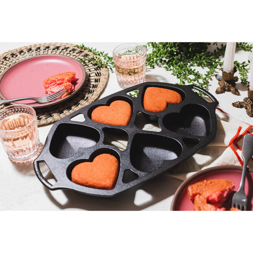 Cast iron heart-shaped mini cake pan with four cavities, featuring three baked red cakes, set on a white tabletop with pink plates, glasses, and brass candle holders, part of Kitchen & Food and Cookware collections.