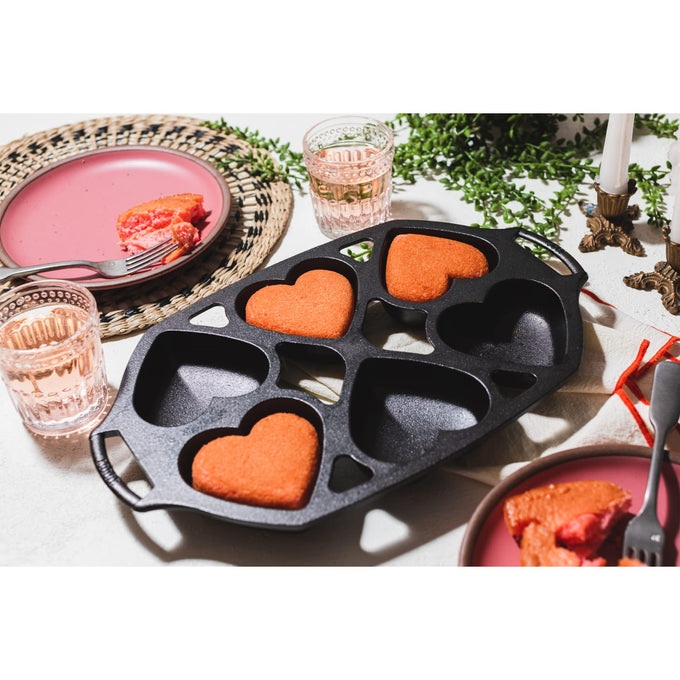 Cast iron heart-shaped mini cake pan with four cavities, featuring three baked red cakes, set on a white tabletop with pink plates, glasses, and brass candle holders, part of Kitchen & Food and Cookware collections.