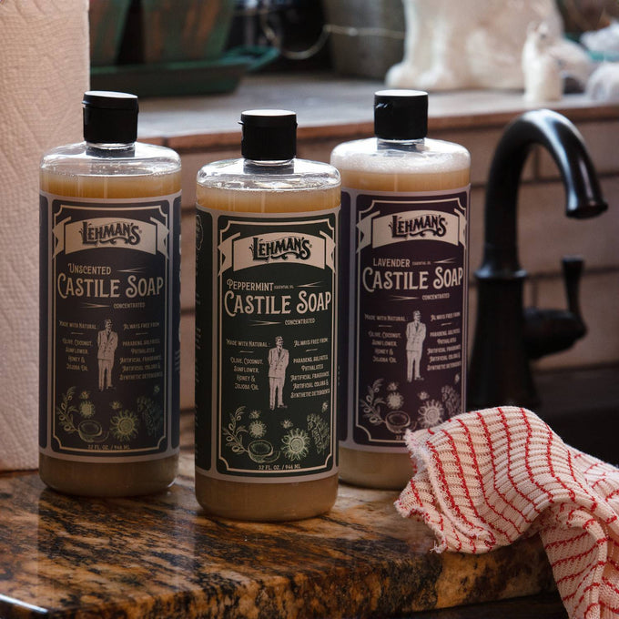 Three handcrafted Lehman's Castile soap bottles in unscented, peppermint, and lavender varieties displayed on a kitchen countertop near a black faucet, highlighting USA made natural liquid soaps for health and grooming collections.
