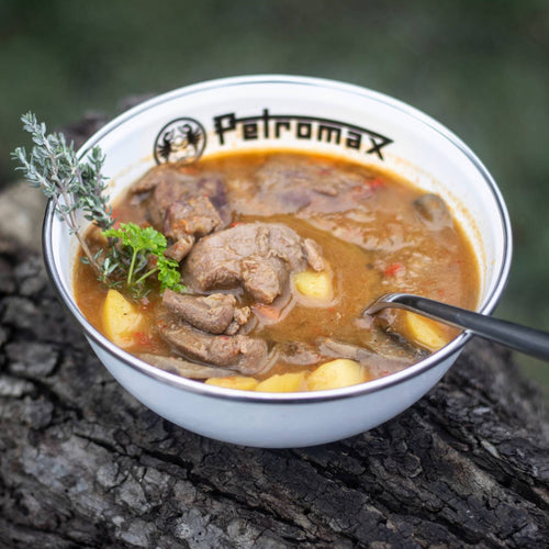 White enamel bowl with black rim and logo filled with hearty beef stew containing potatoes, garnished with fresh herbs, shown outdoors on rough bark surface, suitable for Farm, Garden & Tool, Kitchen & Food, Camping, Dinnerware, Best Sellers, and Promo Eligible collections.