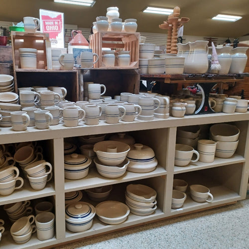 Handcrafted cream-colored stoneware kitchenware including mugs, crocks, and bowls with blue accent lines, displayed on shelves in a rustic food preservation and fermenting shop.