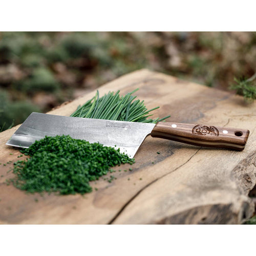 Handcrafted cleaver knife with a hardened steel blade and engraved wooden handle resting on a rustic wooden surface, slicing fresh green herbs for kitchen and camping use.