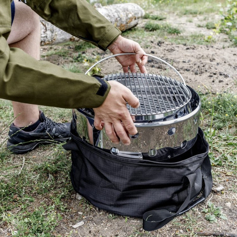 Person placing a portable stainless steel grill with handle into a black ripstop fabric carrying bag outdoors on grass, suited for Farm, Garden & Tool, Camping, Best Sellers, and Promo Eligible collections.