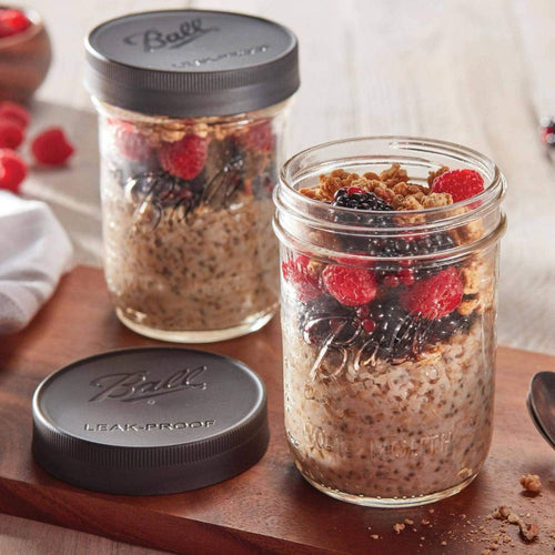 Two wide mouth glass jars filled with overnight oats topped with fresh blackberries, raspberries, and granola, one sealed with a textured dark gray leak-proof lid, set on a wooden board, ideal for Kitchen & Food and Storage and Serving collections.