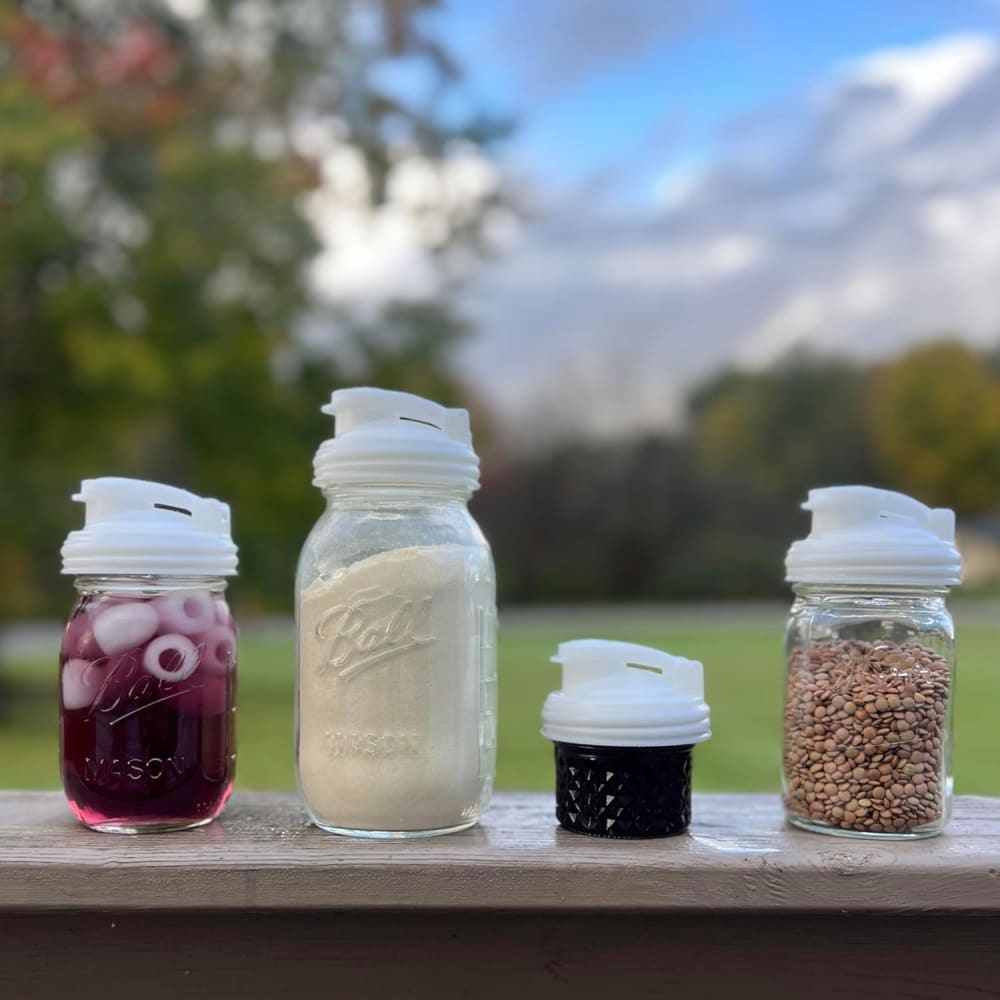 Four clear regular-mouth mason jars and a separate lid with white no-break plastic pour lids featuring flip-top spouts and carry loops, shown outdoors with contents including purple beverage, flour, and lentils, highlighting durable USA-made kitchen accessories perfect for Kitchen Gadgets and Best Sellers collections.