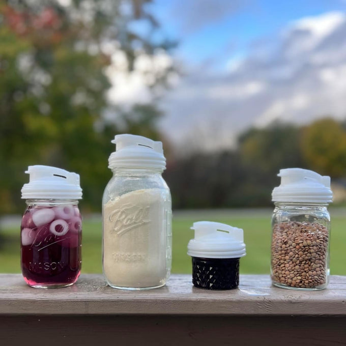 Four clear regular-mouth mason jars and a separate lid with white no-break plastic pour lids featuring flip-top spouts and carry loops, shown outdoors with contents including purple beverage, flour, and lentils, highlighting durable USA-made kitchen accessories perfect for Kitchen Gadgets and Best Sellers collections.