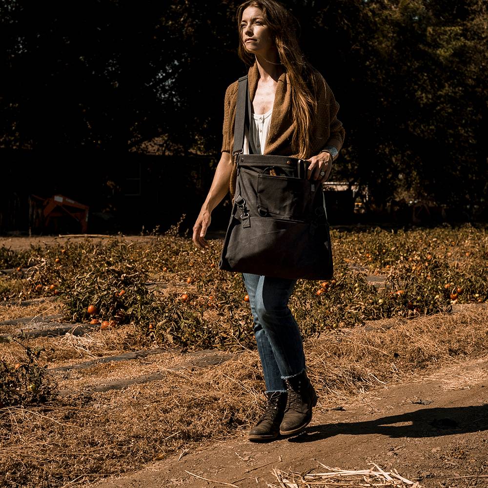 Person walking along a dry orchard path carrying a weather- and water-resistant charcoal gray harvesting bag with convertible straps, ideal for Farm, Garden & Tool and Apple Harvest Supplies collections.