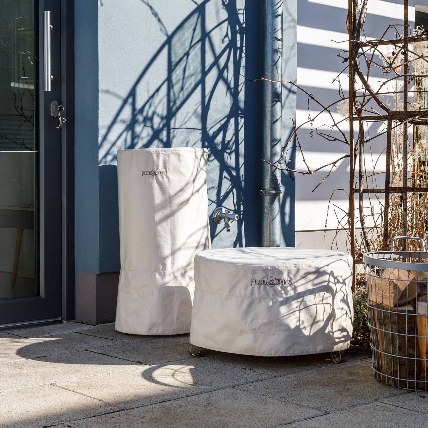 Two beige storage covers made of durable ripstop fabric protecting outdoor fire bowls, shown in a sunny patio setting near a garden hose and wire basket, suited for camping and farm use.