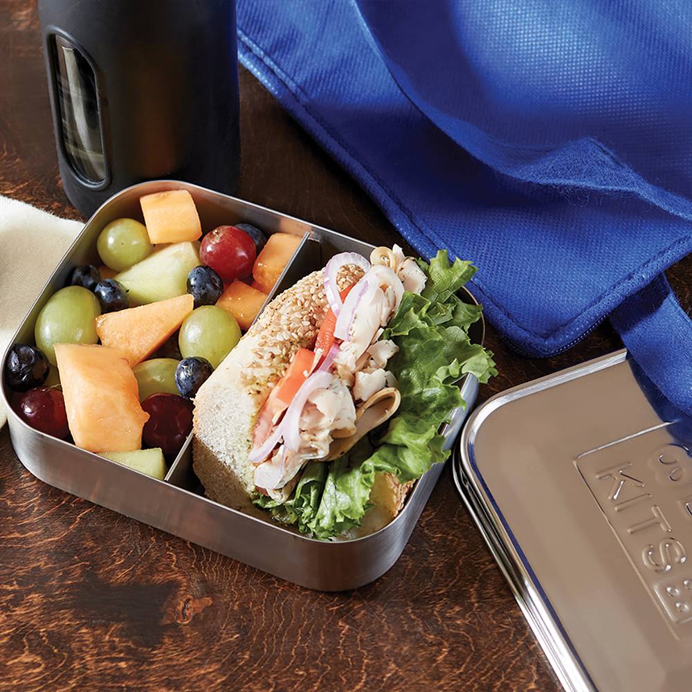 Stainless steel snack container with two compartments holding a fresh sandwich with lettuce and mixed fruit, shown alongside a blue cloth and metal lid, ideal for Kitchen & Food and Storage and Serving collections.