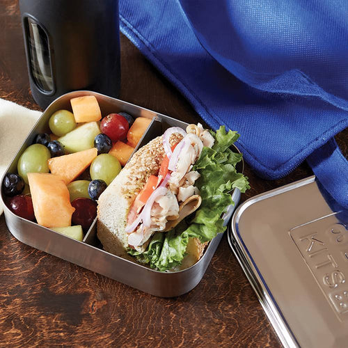 Stainless steel snack container with two compartments holding a fresh sandwich with lettuce and mixed fruit, shown alongside a blue cloth and metal lid, ideal for Kitchen & Food and Storage and Serving collections.