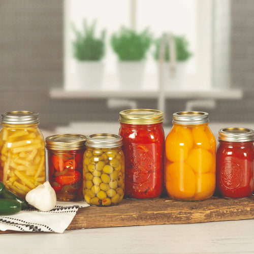Assortment of food-filled glass jars sealed with metal canning lids, featuring a variety of preserved vegetables and fruits, displayed on a rustic wooden board in a kitchen setting