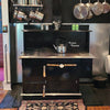 Black vintage-style cookstove with porcelain finish and wood handles, equipped with a kettle on top, surrounded by hanging metal pots and pans, ideal for Stoves, Cookstoves, and Best Sellers collections.