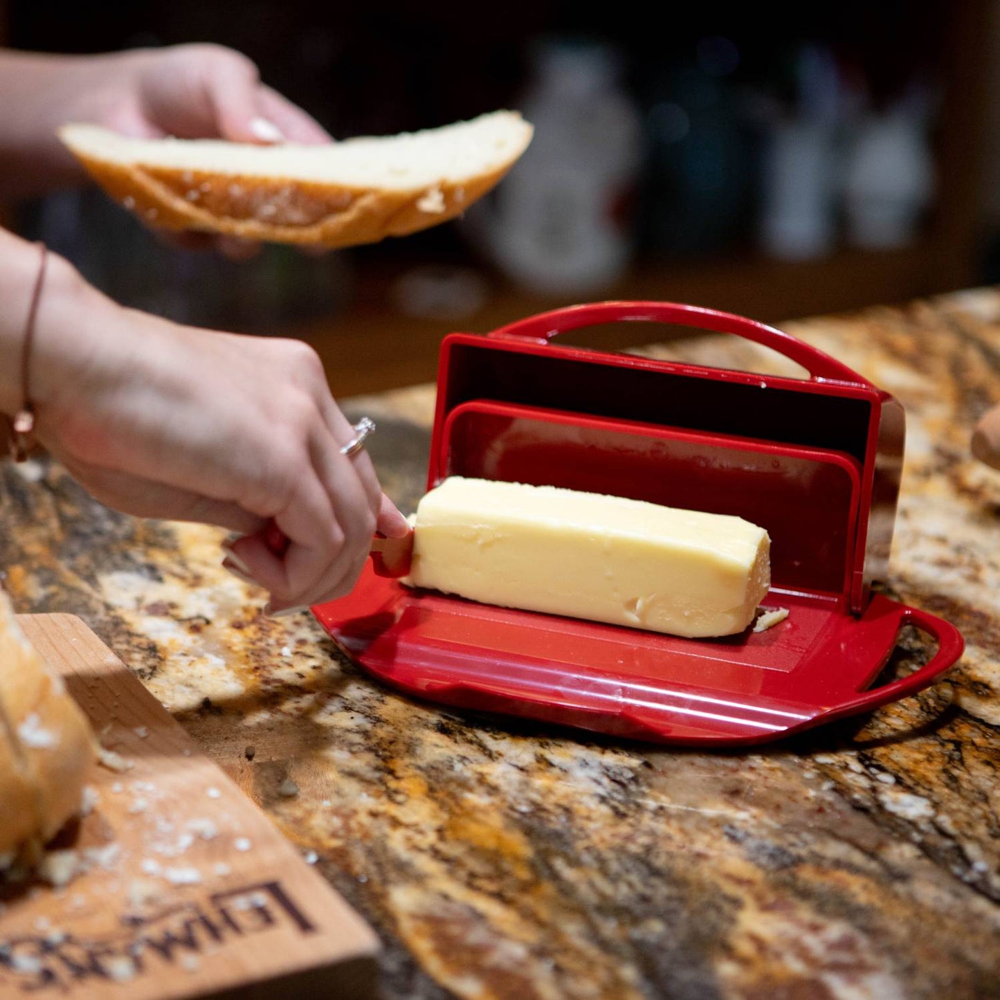 Hand spreading soft butter from a bright red flip-top dish with matching spreader onto a slice of bread on a granite countertop, perfect for Kitchen & Food and Kitchen Accessories collections.