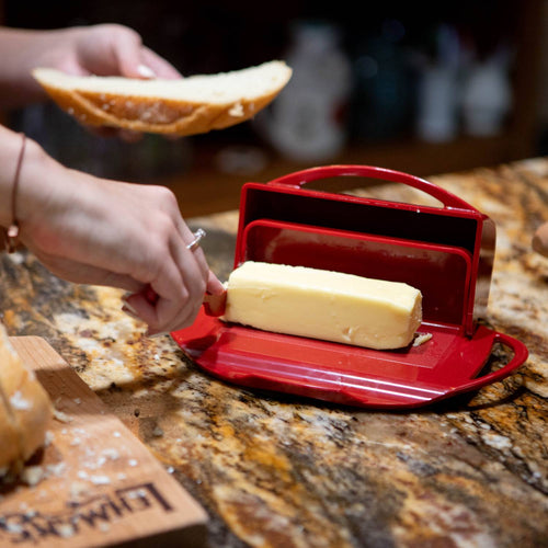 Hand spreading soft butter from a bright red flip-top dish with matching spreader onto a slice of bread on a granite countertop, perfect for Kitchen & Food and Kitchen Accessories collections.
