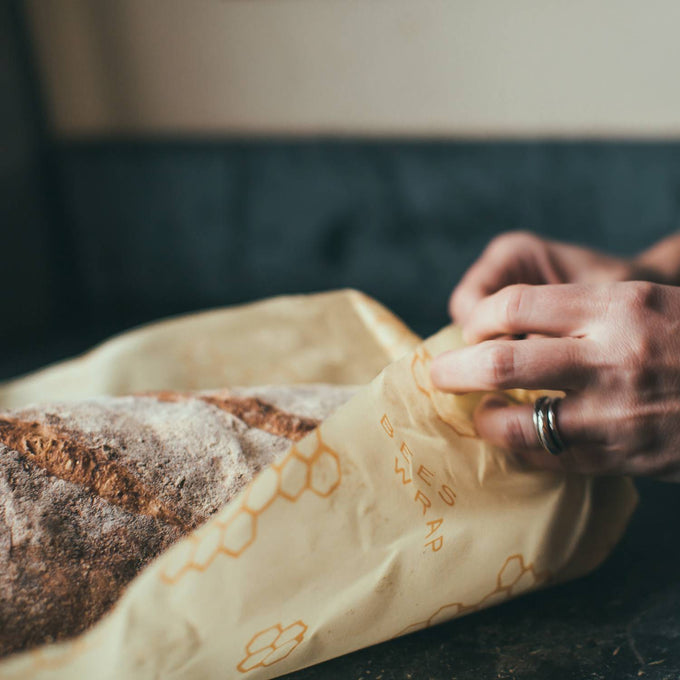 Hands wrapping a rustic loaf of bread with a handmade beeswax food wrap printed with honeycomb designs, part of Kitchen & Food, Storage and Serving, USA Made, and Best Sellers collections.