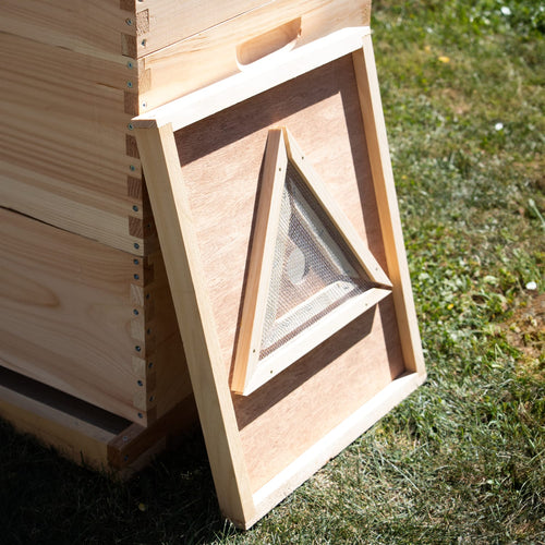Handcrafted wooden bee escape board with a triangle maze design, made in the USA, leaning against a beehive on grass, perfect for chemical-free beekeeping in farm and garden tools.