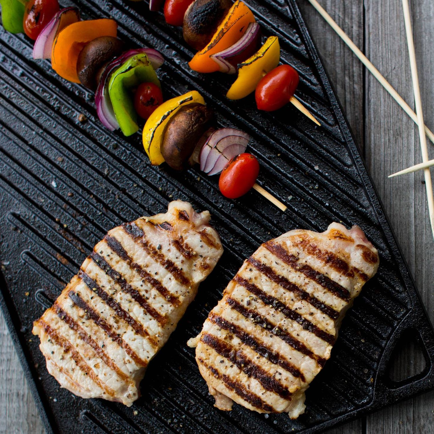 Black cast iron griddle with ribbed surface grilling two seasoned chicken breasts and colorful vegetable skewers, handcrafted USA made cookware for Kitchen & Food and Cast Iron collections.