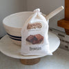 Cloth bag of Lehman's Brownie Mix with a wooden scoop, displayed on a marble cake stand in a kitchen setting, part of Kitchen & Food, Pantry, and USA Made collections.