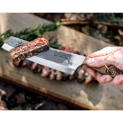 Handcrafted chef’s knife with high-quality steel blade and walnut handle, demonstrating sharpness by slicing a seasoned piece of grilled meat, ideal for outdoor cooking and camping.