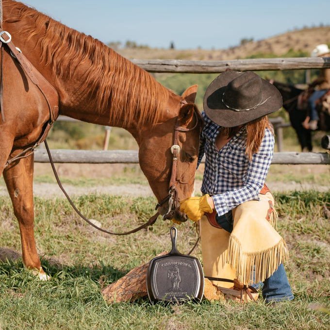 Handcrafted cast iron grill pan with an embossed cowboy design, made in USA, displayed outdoors next to a horse and rider in western attire, fitting Kitchen & Food and Cast Iron collections.