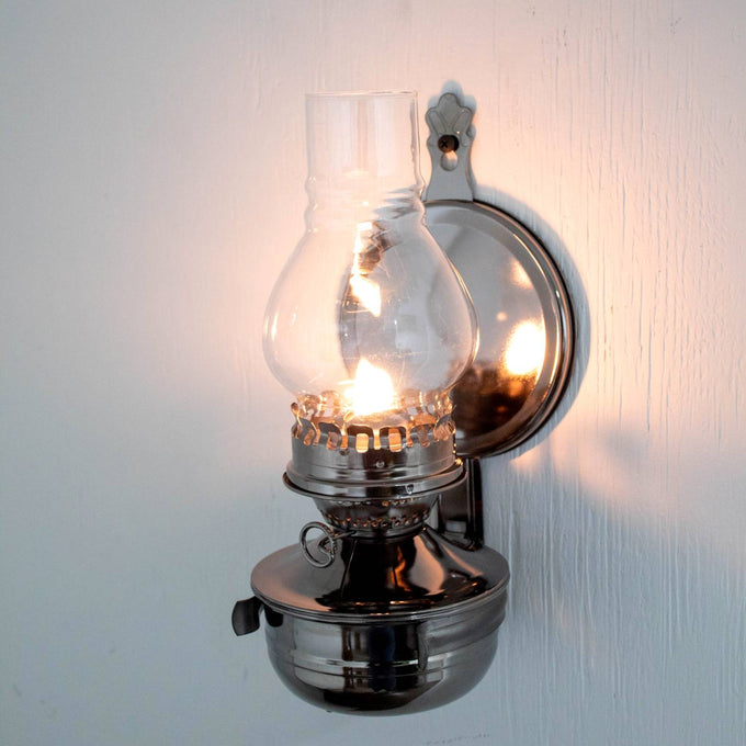 Wall-mounted oil lamp with a painted pewter-like finish, clear ribbed glass chimney, and round reflector casting a warm glow, handcrafted and featured in lamps & lighting and oil lamps collections.