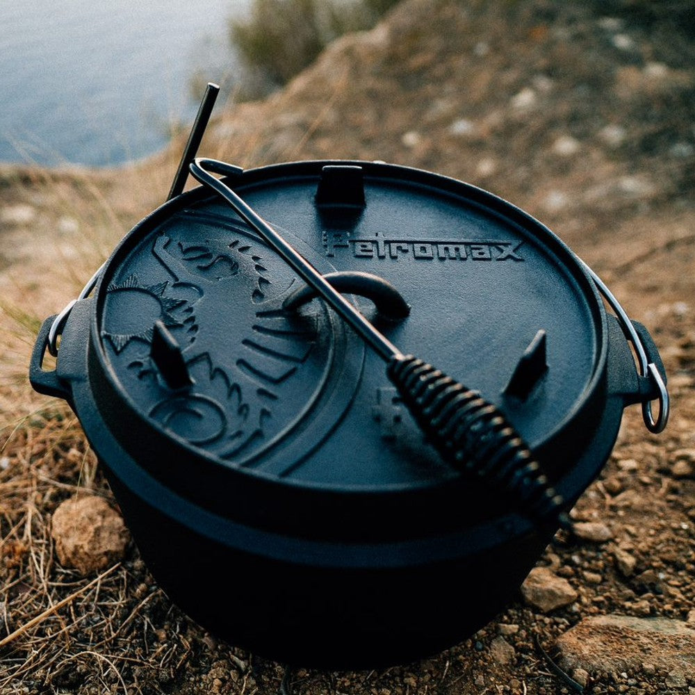Handcrafted cast iron Dutch oven with flat base and embossed dragon lid design, accompanied by a metal handle tool, set outdoors near a lakeside, perfect for Kitchen & Food, Cookware, Cast Iron collections.